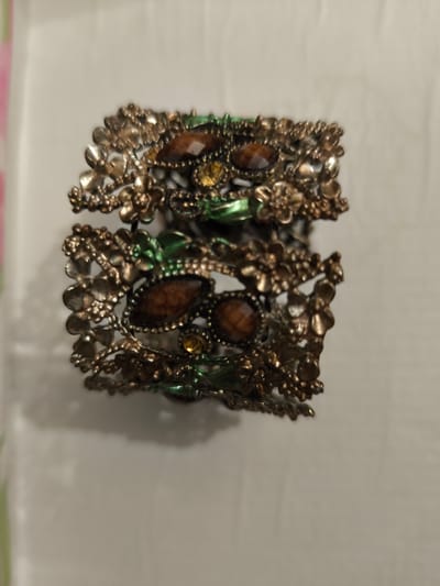 A close-up, slightly blurry shot shows an ornate, bronze-colored metal cuff bracelet with floral motifs and inset brown and yellow gemstones. The bracelet is designed in two tiers, with the upper tier featuring a more prominent arrangement of flowers and gemstones, while the lower tier is more delicate. The background is a plain, light-colored surface, possibly a table or wall, with a soft shadow cast by the bracelet. There is no indication of the bracelet's location in the city of Bolshaya Martynovka, Russia, as the image is an isolated product shot.