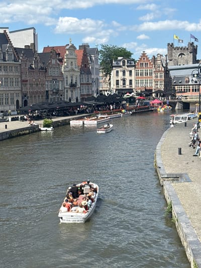 This image depicts a vibrant scene in Ghent, Belgium, with a canal running through the city. The time of day appears to be daytime, with clear blue skies and scattered clouds, suggesting pleasant weather. The architecture along the canal is characteristic of Ghent, featuring historic brick buildings with distinct gabled roofs and large windows.
Several boats are visible on the canal. The most prominent is a white tour boat filled with people enjoying a scenic ride. Passengers are seated, some wearing hats, and appear relaxed and engaged with their surroundings. The boat has visible text on its side: "32 (0)9 / 269 08 69", likely a phone number for boat tours. Another smaller white boat is also present.
Along the canal's edge, there's a cobblestone walkway lined with outdoor seating areas, indicating a popular spot for cafes or restaurants. People are gathered at these tables, some under black umbrellas, suggesting a lively atmosphere. Further along the canal, a procession of colorful flags, including rainbow-colored ones, adds a festive touch to the scene. The overall mood is one of a bustling, enjoyable day in a historic European city.