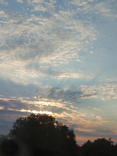 The image captures a sunset over Hadiach, Ukraine. The sky is filled with a mix of small, fluffy clouds and larger, darker formations. Rays of sunlight are piercing through the clouds, creating a dramatic effect. In the foreground, the silhouette of a large tree is visible, with its branches obscuring part of the setting sun. The lower portion of the image is dark and appears to be a landscape, possibly with more trees or buildings, but they are not clearly discernible due to the low light conditions and the focus on the sky. The overall impression is one of a serene and beautiful natural phenomenon.