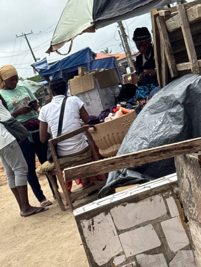 In Ajara, Nigeria, a street vendor is seen tending to her goods. She is wearing a black headscarf and a blue and black patterned outfit. To her left, another woman in a light green shirt and headscarf is looking at her phone. A third person, partially visible on the far left, is wearing a white shirt and grey pants. They are standing in front of a makeshift stall constructed from wood and covered with a tarp. Various items, including boxes and what appear to be bottles of products, are displayed on the stall. The ground is unpaved.