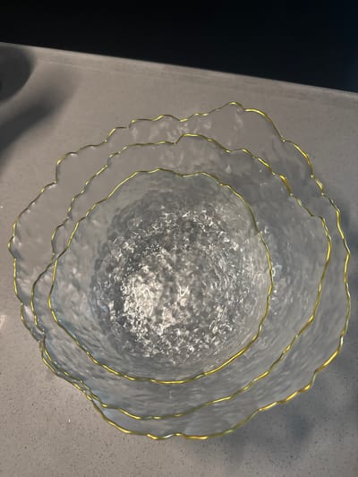 This is a high-angle, close-up, full-shot image of three overlapping, clear, textured glass bowls with ruffled edges and gold trim. The bowls are arranged in a nested formation, with the largest bowl on the outside and the smallest bowl in the center. The surface of the bowls is irregular and bumpy, reflecting light and creating a shimmering effect. The gold trim follows the wavy edges of each bowl, adding a decorative touch. The bowls are placed on a light gray countertop, which has a subtle speckled texture. A dark, out-of-focus area is visible in the upper portion of the image, suggesting an overhead light or background. The scene is indoors, likely in a kitchen or dining area. The location is Shunde District, China.