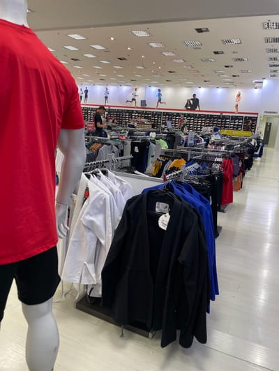 The image captures the interior of a retail sporting goods store located in Anápolis, Brazil. In the immediate foreground, a white male mannequin is visible from the back, wearing a red short-sleeved t-shirt and black shorts. To the right and slightly in front of the mannequin, a clothing rack displays multiple white martial arts uniforms (gis), and another rack holds black and blue martial arts uniforms; a tag on a black uniform reads "VALIJA." The midground features several aisles of clothing racks presenting various activewear garments. Behind these racks, a long display wall holds numerous athletic shoes organized on shelves. Above the shoe display, a large mural depicts five stylized figures engaged in running and stretching. A single male individual wearing a dark shirt is visible in the middle-left, looking down, possibly at a mobile phone. Other indistinct figures are present further in the store. On the far right wall, a partial sign indicates "PROVADORES" (Fitting Rooms). The ceiling is a grid system with numerous square, recessed fluorescent light fixtures. The flooring is a light-colored, reflective tile.