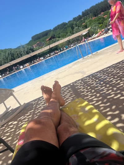 An aerial perspective shows a person's legs and feet resting on a yellow towel, positioned on a tiled patio next to a large outdoor swimming pool. The pool is filled with bright blue water, and several individuals are visible near its edge and within the water. A woman in a pink cover-up and flip-flops stands to the right of the frame, facing away from the camera. The area is shaded by a patterned canopy, casting a dappled light across the patio. In the background, a lush green hillside is dotted with houses and trees. The sky above is a clear, vibrant blue. This scene appears to be at a resort or public swimming facility located in Delchevo, North Macedonia.