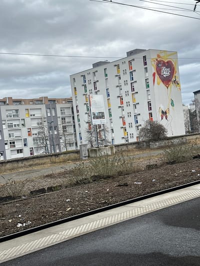 The image displays an urban scene in Saint-Louis, France, captured from the perspective of a train platform, looking towards residential buildings under an overcast sky.

In the foreground, a dark grey asphalt train platform with a textured light grey tactile paving strip runs along the bottom edge. Immediately adjacent to the platform are railway tracks, consisting of dark grey ballast stones, visible wooden sleepers, and metal rails. Scattered white debris, resembling paper or plastic, is distributed across the ballast. Sparse, dry, brownish-green vegetation grows in clusters among the tracks.

The midground features a low, aged concrete retaining wall, beyond which the ground level appears elevated. Behind this wall, a prominent, multi-story residential building with a bright white facade stands on the right. This building's windows are framed in various vibrant colors including red, orange, yellow, green, blue, and purple. The right gable wall of this building is adorned with a large mural depicting a stylized red and orange heart with yellow accents. Within the heart, indistinct text, possibly "SAUTONS L'EMPRISE," is visible. Two bees are painted near the heart, and below it, a large brown-orange butterfly and a smaller green butterfly are connected by a thin line descending from the heart. Directly in front of this building, a white rectangular sign on a pole displays the letters "BTSC" at the top, accompanied by a logo, and smaller text and imagery below, appearing to advertise a business school. To the left, another multi-story residential building with a lighter grey facade and a mix of colorful window frames and white roller shutters is partially visible. Bare deciduous trees with dark branches are situated between the buildings and the retaining wall, alongside some dense brown shrubbery.

The background consists of a uniformly light grey, overcast sky. Thin black lines, indicative of overhead power lines or catenary wires, traverse the upper portion of the frame horizontally. Additional distant buildings are partially visible. No individuals or discernible human activity are present within the scene.