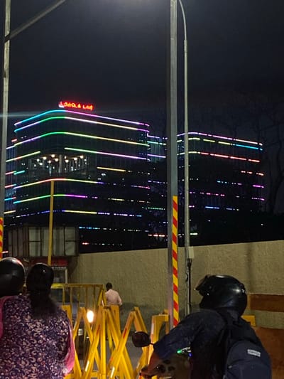 The image depicts a nighttime scene in Chennai, India, dominated by a brightly lit, modern building adorned with vertical lines of colorful LED lights. The building's facade reflects these lights, creating a vibrant and dynamic visual. In the foreground, a person wearing a helmet and dark clothing is visible, appearing to be on a yellow scissor lift or similar elevated platform. Another person, wearing a purple and white patterned garment, is also partially visible on the platform.
The activity suggests some form of maintenance, installation, or inspection work being conducted on or around the building at night. The presence of the scissor lift and the people on it points to an operational task.
Notable details include the time of day being night, indicated by the dark sky and artificial lighting. The bright, colorful lights on the building suggest a festive or modern urban environment. The visible text in Tamil on the building, which appears to be "பிளாட்டினம் டவர்" (Platinum Tower), provides a specific location cue. The yellow and red striped barrier on the scissor lift is a common safety feature. The overall mood is one of urban activity and illumination, despite the late hour.
