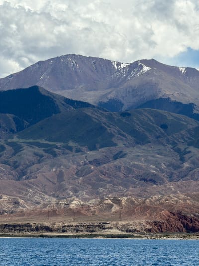 The image shows a mountainous landscape in Tong, Kyrgyzstan, next to a large body of water. The mountains exhibit varying shades of brown and grey, with some snow visible near the peak of the highest mountain on the left. Dense vegetation covers a significant portion of the mountains. The water, on the right side of the image, is a deep blue with a slight ripple texture. The sky is cloudy, with white clouds partially obscuring the mountain peaks.