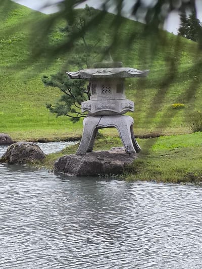 A stone lantern sits on a rock formation beside a body of water. The lantern is traditional in design with intricate carvings. The water is rippling, suggesting a light breeze. The background consists of lush green grass and trees. The overall scene appears to be a serene outdoor setting, possibly a park or garden in Mažučiai, Lithuania.