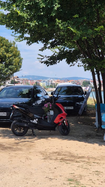 The image depicts a street scene in Ravda, Bulgaria, featuring two black cars and a red scooter parked on a sandy area under trees. The scooter is in the foreground, positioned between the two cars. The first car, an Audi, is on the left, with its license plate partially visible as "03 PH". The second car, a BMW, is on the right, with its license plate reading "3832 PH". Behind the vehicles, a town with colorful buildings and a body of water can be seen. The sky is blue with some clouds. The overall environment suggests a sunny day in a coastal area.