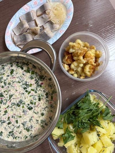 The image shows a top-down view of a wooden table laden with several dishes of food. In the upper left, a plate contains sliced pickled herring arranged on a decorative plate with a floral pattern. To the right of the herring is a clear plastic container filled with small, golden-brown fried pieces, possibly cauliflower or potatoes. In the center left, a large metal pot is filled with a creamy soup or cold dish containing finely chopped green herbs and possibly red onion. In the lower right, a glass dish holds cooked, cubed potatoes garnished with fresh parsley and dill. The scene appears to be a home-cooked meal. The location is not specified in the image.