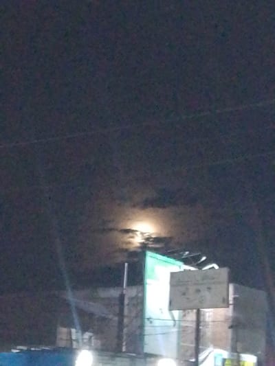 The image depicts a night scene in Gampongpaya, Indonesia. The sky is dark and cloudy, with a bright moon partially obscured by clouds, casting an ethereal glow. In the foreground, there are structures that appear to be buildings or market stalls, some illuminated with artificial light. A signpost with a white board is visible, likely displaying information or advertisements. The overall impression is a quiet, possibly bustling, night in an urban or semi-urban area.