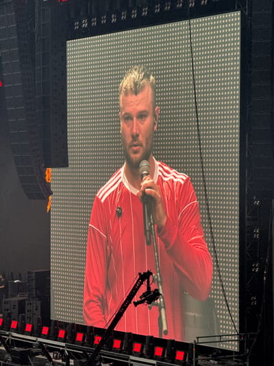 The image depicts a large screen displaying a close-up of a male performer on a stage, likely during a concert in Dusseldorf, Germany. The performer is wearing a red long-sleeved shirt with white stripes on the shoulders and is holding a microphone. He has short, blond hair and appears to be singing or speaking into the microphone. The screen is illuminated with a grid of white dots. To the left and right of the screen, large speaker stacks are visible, suggesting a large venue. In the foreground, stage lighting equipment and a camera crane are present. The overall environment appears to be a concert hall or stadium setup.