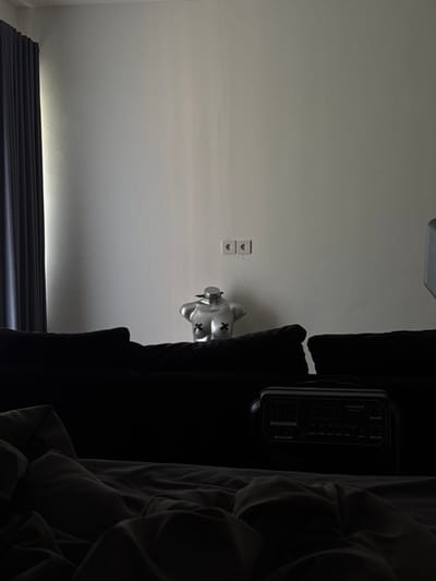 The scene is an indoor room in Beraban, Indonesia, characterized by subdued natural lighting primarily from the left. A light-colored wall, exhibiting multiple fine cracks, occupies the background. Two white electrical outlets are horizontally mounted on the wall, slightly right of center. To the far left, dark, pleated curtains are drawn, obscuring a window. In the midground, a silver, metallic, headless female torso mannequin, adorned with black "X" marks over the nipples, is positioned behind a dark-colored sofa. The upper portion of a black portable power station, branded "GOLABS" and featuring a display and control panel, rests on or immediately behind the sofa in the lower right. The immediate foreground is dominated by dark, rumpled bedding, suggesting a bed or a sofa with covers. No people are present.