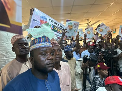The image shows a crowded room, presumably indoors, filled with people holding signs and promotional materials for "YB IKARA" for Senate Zone 2027. Many individuals are wearing traditional caps. There are numerous cameras and recording equipment, indicating a press conference or public event. The environment seems to be a well-lit, possibly public, space. No specific details indicate Shaba, Nigeria.