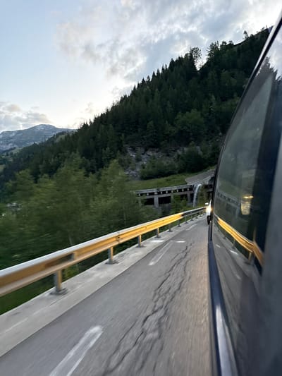 This is a subjective shot from a moving vehicle on a paved road in a mountainous region, likely near Sainte-Foy-Tarentaise, France, as indicated by the presence of evergreen trees on a steep slope and a distant, snow-capped mountain peak under a cloudy sky. The perspective is low and angled, showing the right side of the road with a yellow guardrail. The road itself has visible cracks and white markings. To the right, the dark-colored side of a vehicle, possibly a car or van, takes up a significant portion of the frame, reflecting the sky and surrounding environment. In the background, a concrete structure, possibly an aqueduct or bridge, with arches and a cascading stream of water, is visible on the mountainside. The overall impression is one of travel through a scenic, natural landscape.