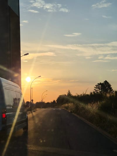 The image shows a street scene at sunset in Leudelange, Luxembourg. The sun, a bright orb with prominent lens flare, is low on the horizon, casting a warm, golden light across the sky and the road. The sky is a gradient of soft orange and yellow near the sun, transitioning to a pale blue at the top, with scattered white clouds.
On the left side of the road, a large, dark, industrial-looking building looms, its textured facade catching some of the sunlight. A silver van is parked or driving on the left side of the road, its rear visible in the foreground. The van's brake light is illuminated. Streetlights with curved poles are visible along the roadside, some illuminated and others not.
The road itself appears to be asphalt, with some texture and markings visible. On the right side of the road, a dense line of vegetation, primarily tall grasses and bushes, runs along the embankment. A few trees, including a prominent evergreen, are silhouetted against the sunset sky. Sunlight streams through the grasses, creating a bright, almost ethereal effect. The overall impression is one of a tranquil, late-day rural or semi-urban setting.