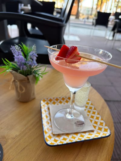 The image displays a refreshing drink arrangement and decorative elements set on a light wooden table, suggesting a relaxed atmosphere in an indoor venue like a cafe or restaurant.

Centered in the frame is a stemmed cocktail glass, resembling a martini or margarita glass, filled with a frosty, pale pink beverage. Condensation is visible on the glass's surface, indicating a cold drink. Two vibrant red strawberry slices are skewered on a wooden stick, serving as a garnish. Adjacent to the cocktail, a smaller, slender glass, possibly a shot glass or mini champagne flute, holds a clear or very pale yellow, bubbly liquid, likely sparkling wine or a mixer.

Both glasses are placed on a small, rectangular ceramic tray. The tray features a distinctive pattern of interlocking yellow geometric shapes, similar to quatrefoils, set against a white background, and has a thin blue rim. A light-colored napkin rests beneath the glasses on the tray.

To the left of the drinks, a small bouquet of artificial lavender-like flowers with green foliage is presented in a rustic brown paper or burlap wrap. The table itself is made of light-colored wood, exhibiting visible grain patterns.

The background is softly blurred, showing bright areas and reflections consistent with windows or a well-lit interior, suggesting it is daytime. No individuals are present in the image. A faint, circular, unreadable logo or text is barely discernible on the base of the main stemmed glass. The overall scene evokes a sense of leisure and enjoyment, fitting for a relaxed moment in a location such as Salou, Spain.