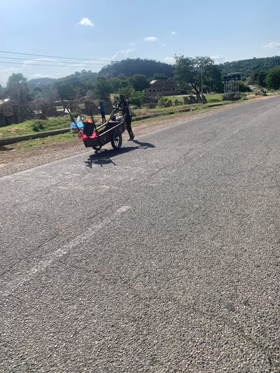 A person is pushing a bicycle with a trailer attached. The trailer is laden with bags and other items. The person is wearing a black shirt and shorts, and a helmet. The bicycle and trailer are on a paved road. In the background, there are trees, hills, and some buildings. The sky is blue with a few white clouds. The sun is shining, suggesting it is daytime. The scene appears to be rural, possibly in Boi A, Nigeria, as indicated by the location context. There are no visible signs of text in the image. The overall mood is one of travel or movement, likely a person transporting belongings.