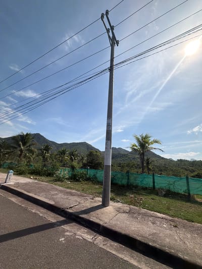 The image captures a roadside scene in Vĩnh Lương, Vietnam, under a bright, partly cloudy sky. A concrete utility pole, adorned with multiple wires and insulators, stands prominently in the foreground, casting a shadow on the adjacent pavement. To the left, a paved road runs alongside a raised concrete sidewalk with a curb. A small, blue and white marker post with the inscription "H17 2" is visible on the sidewalk.
Beyond the sidewalk, a stretch of green grass and vegetation is bordered by a vibrant green mesh fence. In the background, lush, tree-covered mountains form a picturesque backdrop. Several palm trees, characteristic of the tropical climate, are scattered throughout the landscape, adding to the scenic beauty. The sun is high in the sky, with its rays creating a lens flare effect, indicating a clear and sunny day. The overall impression is one of a rural or semi-rural area, possibly on the outskirts of Vĩnh Lương, with infrastructure for electricity and a natural mountainous environment.