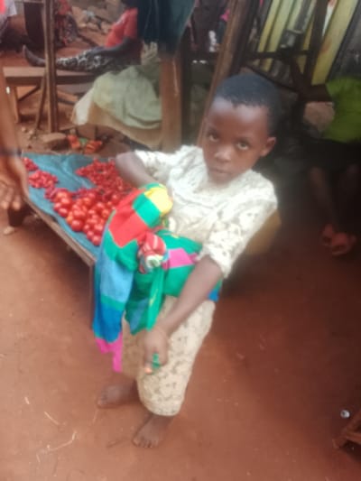 This image is a medium shot depicting a young child, likely a girl, standing in what appears to be an outdoor market or roadside stall. The child is centrally positioned, looking directly at the camera with a serious or thoughtful expression. She has dark skin and short, neatly cut dark hair.
The child is wearing a light-colored, possibly cream or white, long-sleeved top with a delicate, lacy, or textured pattern. Her lower garment is a light-colored skirt or wrap with a subtle, possibly floral or abstract, pattern, extending below her knees. Around her waist, she has a brightly colored piece of fabric, possibly a wrap or a child carrier, featuring prominent blocks and patterns in red, blue, green, yellow, and pink. Her bare feet are visible, placed on the reddish-brown dirt ground. A thin, light-colored bracelet can be seen on her right wrist.
To the child's left, there is a wooden table or low stand covered with a bright blue tarp. Piled high on this tarp are numerous fresh red tomatoes and smaller, elongated red peppers (or small, pointed tomatoes), suggesting a produce stall. A person's arm and hand are partially visible on the far left of the frame, near the table, indicating another individual is nearby, possibly tending the stall.
The background is somewhat blurred but provides context. Wooden structures, possibly parts of market stalls or simple furniture, are visible. On the right side of the image, further back, a person can be seen sitting on a wooden chair, wearing a green garment. Below them, another person's legs and feet, wearing pink open-toed slippers, are partially visible. The ground throughout the scene is a consistent reddish-brown, characteristic of dirt or earth. The lighting suggests it is daytime, with no harsh shadows, implying either an overcast day or an area with some overhead cover. There is no visible text in the image.
