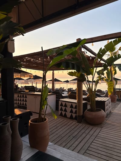 This image captures a serene sunset scene at a beach bar located in the Pallini Municipal Unit of Greece. The foreground features a wooden deck with several large, textured terracotta vases and planters. In the middle ground, a bar area is visible with white cabinets decorated with black triangular patterns, and various seating arrangements with cushions and umbrellas are set up on the sand, facing the sea. Lush green plants, likely banana trees, are prominently displayed in large pots, adding a tropical ambiance to the setting. The background reveals the calm sea bathed in the warm glow of the setting sun, with faint outlines of distant landmasses or islands. The sky transitions from a soft orange hue near the horizon to a pale blue overhead. String lights are draped across the thatched roof of the bar area, suggesting it is prepared for evening service. A single person is partially visible behind the bar, and another figure is seated on the beach in the distance. The overall atmosphere is one of peaceful relaxation and natural beauty.