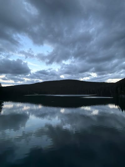 The image displays a serene landscape in Pitomine, Montenegro, characterized by a large body of water (likely a lake) under an overcast sky, framed by dark forested hills. The upper half of the frame is dominated by a heavy, textured cloud cover, predominantly dark gray and bluish-gray, with several lighter patches of pale blue sky visible, particularly towards the center-left and along the horizon. Below the sky, a dense, dark green forest covers undulating hills or mountains, forming a continuous silhouette across the middle ground. The lower half of the image features the surface of the lake, which exhibits distinct reflections of the clouds and sky. The water's surface is not entirely still, showing subtle ripples and undulations that slightly distort the reflected patterns. The overall lighting is dim, suggesting either dawn, dusk, or heavily overcast daytime conditions. No people or artificial structures are visible in the scene.
