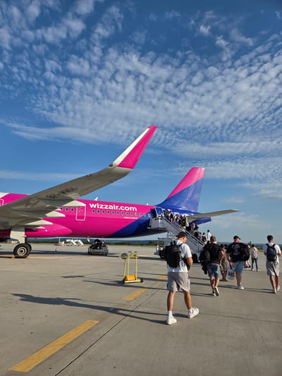 The image shows a Wizz Air airplane on a tarmac. Several people are boarding the plane using a set of stairs. The airplane is predominantly pink and blue, with "wizzair.com" printed on the fuselage. The sky is blue with scattered white clouds, suggesting it is daytime with fair weather. The ground is paved, with yellow lines indicating parking or taxiing areas. In the background, there are some trees and buildings, possibly indicating an airport environment. The people appear to be passengers, dressed in casual clothing. Some are carrying backpacks. Their body language suggests they are in the process of boarding or waiting to board the aircraft. The overall scene depicts a typical airport boarding operation on a clear day. The visible text on the aircraft is "wizzair.com" and "AIRBUS A321 NEO". The registration number of the aircraft is partially visible as "HA-M...", likely "HA-MHM". The location context provided is Aksakovo, Bulgaria.