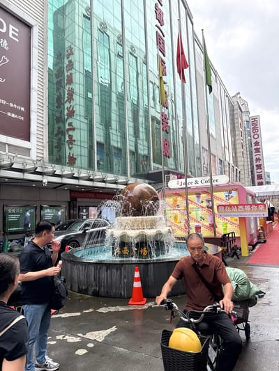 In Yuexiu District, China, a man with graying hair, wearing a brown polo shirt and black pants, rides an electric bicycle. The bicycle has a black basket on the front containing a yellow helmet and a green bag is strapped to the rear rack, obscuring the bicycle's frame. Behind him, a large, circular fountain is active, with water spraying upwards from a brown spherical object at its center. An orange traffic cone is placed to the right of the fountain. In the foreground, to the left, two individuals stand facing the fountain; one, a man in a black polo shirt and blue jeans, holds an ice cream cone, while the other, a woman in a black shirt, is partially visible. The background features a modern building with a glass facade reflecting the sky and surrounding structures. A pink food stall with colorful images of drinks and Chinese text is situated near the fountain. A red carpet extends along the right side of the scene.