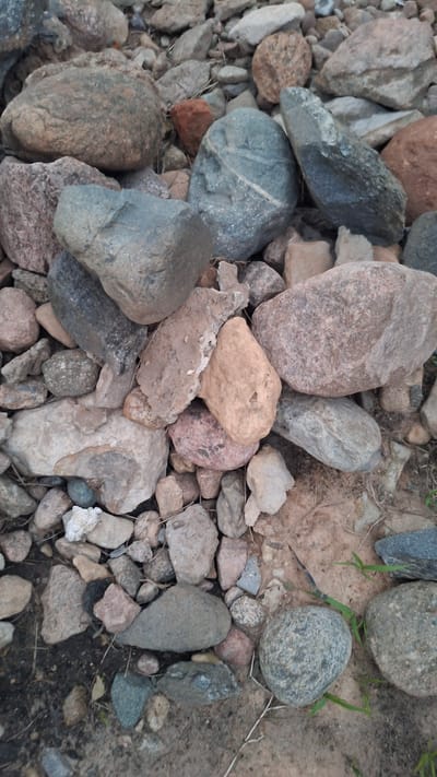 The image displays a close-up, overhead view of a variety of stones and pebbles scattered on the ground. The stones vary in size, shape, and color, with hues of gray, brown, tan, and reddish-brown. Some stones are smooth and rounded, while others are jagged and angular. The ground beneath the stones appears to be a mixture of dirt and sand, with some sparse vegetation, likely grass and dried twigs. There are no people or animals visible in the image. The lighting suggests it might be daytime, with the overcast sky possibly diffusing the light. The scene is a natural environment, possibly a riverbed, gravel path, or area with exposed soil and rocks. It is not possible to determine the specific location within the city of Stowbtsy, Belarus, from the image itself.