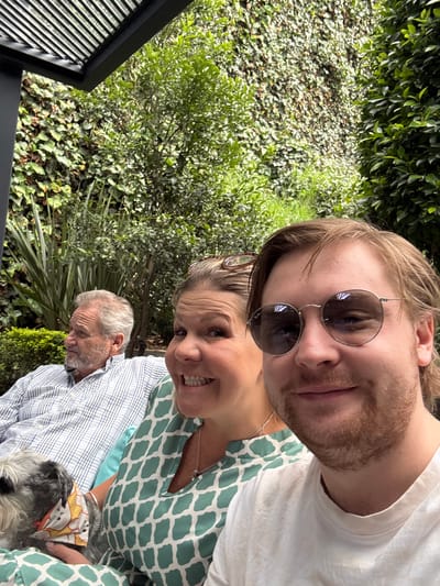 The image shows three people and a dog on a covered patio. A man with gray hair and a beard is seated on the left, wearing a blue and white plaid shirt. In the center, a woman with blonde hair is smiling and looking towards the camera, wearing a green and white patterned dress. On the right, a young man with blonde hair, wearing sunglasses and a white t-shirt, is looking at the camera. A small gray dog with a bandana is being held by the woman. The background consists of lush green foliage and a vine-covered wall. The scene appears to be in a garden or outdoor seating area, possibly in Huixquilucan de Degollado, Mexico, given the commonality of such upscale outdoor living spaces in the region.