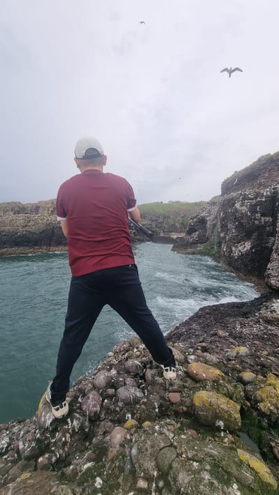 A person, viewed from behind, stands on a rocky outcrop overlooking a body of water. The person is wearing a maroon t-shirt, dark pants, a white baseball cap, and light-colored sneakers. They are holding a black object, possibly a fishing rod, horizontally. Several birds are visible in the sky. The environment consists of rugged, rocky cliffs and the sea, with some vegetation visible in the distance. The sky is overcast. The scene appears to be outdoors and near the coast, possibly in a natural setting. The location is identified as Mill of Uras, United Kingdom.