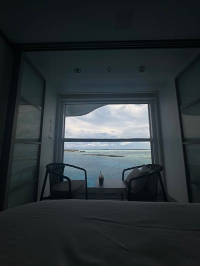 The image displays an interior view of a room, likely a ship cabin, facing a large window overlooking the ocean and land formations of the Berry Islands, Bahamas. In the immediate foreground, a bed with white linens is visible, slightly out of focus. Two dark-framed chairs with light cushions flank a small, light-colored rectangular table positioned directly in front of the window. A single clear cup containing a dark beverage and a straw rests on the table. The window frames an expansive view of multi-toned blue and turquoise ocean water under a partly cloudy sky. In the mid-distance, several low-lying landmasses or cays are visible, with one larger landmass on the left showing structures, possibly a dock, and a tall, slender upright object. A linear formation, possibly a breakwater or reef, extends across the water in the middle distance. The interior of the room is dimly lit, with white walls and ceiling featuring recessed lighting and a ventilation grate. No individuals are present in the scene.