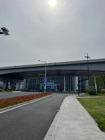 The image shows a street in Hang Zhou Shi, China, located at coordinates 30.273 latitude, 120.419 longitude. The scene is under a bright, sunny sky with no clouds. A large, modern, glass-fronted building stands in the middle of the road. The building has a sign that reads "AUTO" and Chinese text, indicating it is a car dealership. The street is asphalt and has a pedestrian walkway with a sidewalk on the right. Several people are walking in the street and several cars are parked in the distance. The street has a few streetlights and a large, concrete overpass. Trees and green grass are on the right and a bed of red flowers borders the left side of the street. There is a security camera on a pole on the left side of the image.