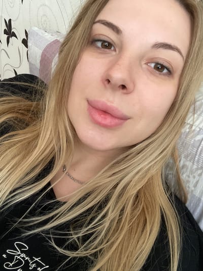 This is a close-up, overhead selfie of a young woman with long, blonde hair. She is lying down, with her head tilted slightly to the side, and her blonde hair is spread across her shoulders and chest. She is wearing a black t-shirt with white text on it. The background appears to be a bed with patterned sheets and a pillow. Her face is the main focus, with her brown eyes looking towards the camera. She has natural-looking makeup, with slightly defined eyebrows and a subtle pink lip color. Her expression is neutral, with a slight, gentle smile. The lighting suggests it could be daytime, likely indoors. There is some visible text on her t-shirt, which appears to be part of a phrase or quote written in cursive. The image is cropped to focus on her face and upper torso, with the background slightly blurred.
