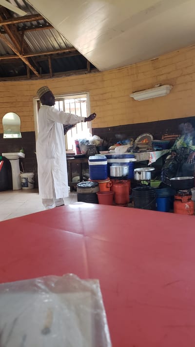 The image depicts a man wearing a white robe and a patterned cap, standing with his arms raised as if in prayer or supplication. He is positioned in what appears to be an indoor space, possibly a workshop or storage area, given the collection of items behind him. The background is filled with various objects, including stacked gas cylinders, pots, coolers, and other containers. A red table is prominently visible in the foreground, partially obscuring the scene. The walls are made of yellow brick, and a window with horizontal blinds is visible on the right. Below the window, a toilet and a bucket are situated on the floor. The overall impression is of a utilitarian or storage space, possibly in a community setting. The man's posture suggests a moment of devotion or deep thought. The lighting appears to be from artificial sources, though a hint of natural light might be coming from the window. No text is discernible in the image. The image is oriented sideways, with the man facing towards the left of the frame.