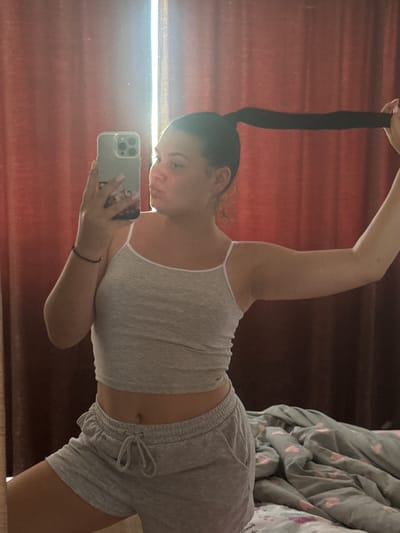 The image shows a young woman taking a selfie in a mirror. She is wearing a grey tank top and grey shorts. Her hair is pulled up in a high ponytail. The room appears to be a bedroom, with a bed and blankets visible in the background. The lighting suggests it is daytime, possibly morning or afternoon. The woman's expression is playful, with her lips pursed. The setting includes a red curtain, possibly indicating a window or a wall covering. There are no visible text elements in the image. The image seems to be taken in Rogosh, Bulgaria.