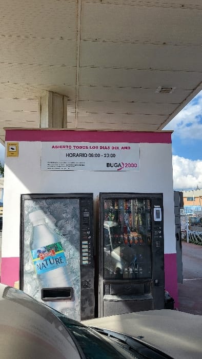 The image displays an outdoor vending machine station situated in Polígon Industrial De Calvià, Spain. A white paneled canopy, showing signs of wear and dirt, extends over a white wall accented with a prominent pink stripe along its top and right side. Two dark-colored vending machines are positioned against this wall. The left machine prominently features a large bottle of Pascual Nature water on its display, while the right machine showcases various packaged snacks and beverages through its transparent front. Both machines are equipped with payment interfaces and product retrieval slots.
Above the vending machines, a white sign on the wall provides operational information in Spanish: "ABIERTO TODOS LOS DIAS DEL AÑO" (Open every day of the year) and "HORARIO 06:00 - 23:00" (Hours 06:00 - 23:00). The sign also includes the logo "BUGA 2000" and a small security camera icon on the left. The hood and windshield of a light-colored vehicle, possibly silver, are visible in the immediate foreground, partially obscuring the lower sections of the vending machines. The scene is under natural daylight, casting shadows to the left side of the wall. In the upper right background, a blue sky with scattered white clouds is visible, along with distant industrial structures and utility poles. No individuals are present in the scene, and no actions or interactions are depicted.