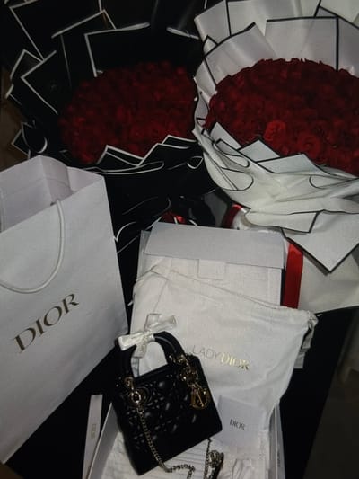 The image depicts a luxurious arrangement of Dior items and red roses, suggesting a special occasion. In the foreground, a small black Dior handbag with a quilted pattern and gold hardware is prominently displayed. The bag is adorned with a white Dior ribbon. To the left of the handbag is a white Dior shopping bag with the brand name in gold lettering. Behind these items, two large bouquets of red roses are arranged with black and white floral paper. Further back, more Dior packaging is visible, including what appears to be a white box and a dust bag with "LADY DIOR" embossed in gold. The overall setting is dark, with the objects illuminated, creating a sense of opulence. The image does not provide any contextual information about the city of Iquique, Chile.