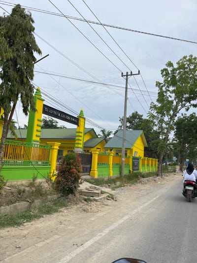 In Bundar, Indonesia, a school building with bright yellow walls and green roofs is visible, bordered by a green and yellow fence. The building has a sign that reads "TK ADHYAKSA XLIV ACEH TAMIANG." A person wearing a white hijab and top is riding a motorcycle away from the camera on a paved road in front of the school. The sky is overcast, and power lines are visible overhead. The surrounding area appears to be a mix of trees and open land.
