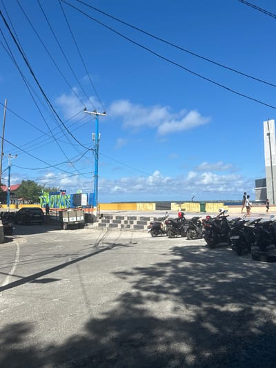 In Bira, Indonesia, a paved area is filled with parked motorcycles under a clear blue sky with scattered clouds. A blue utility pole with numerous wires stands prominently, casting shadows on the ground. In the background, a yellow wall borders the sea, and a modern, tall structure, possibly a monument or part of a building, is visible on the right. Several people are walking near the water's edge. To the left, a black car is parked near a sign with colorful lettering, and a small utility trailer is situated nearby. The scene depicts a bustling outdoor area with elements of urban infrastructure and coastal landscape.