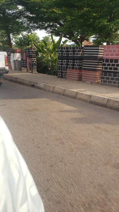 The image depicts a scene in Hayin Banki, Nigeria. Along the edge of a paved road, stacks of red and black patterned bricks are arranged next to the curb. Vegetation, including trees and banana plants, grows behind the bricks. A portion of a white vehicle is visible in the lower left of the frame. The road surface is textured and reflects sunlight.