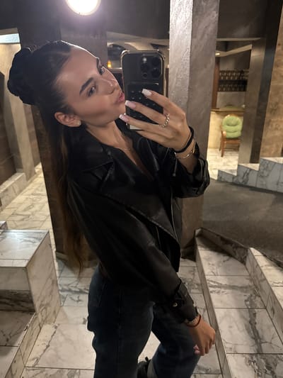 The image is a vertically oriented, low-angle selfie taken by a young woman with long, dark hair and fair skin. She is wearing a black leather jacket, blue jeans, and several pieces of jewelry: a gold bracelet on her left wrist, a silver ring on her right index finger, and a red string bracelet on her right wrist. She is holding a smartphone in her left hand, with her right hand resting on her lap. Her gaze is directed towards the phone's camera.
The background appears to be an indoor staircase with marble-tiled steps and concrete railings. The lighting is dim, suggesting it might be evening or an indoor setting with artificial light. The overall mood is casual and relaxed. No text is visible in the image. The specific location cues suggest Vidin, Bulgaria, but this is not visually evident from the image content itself.