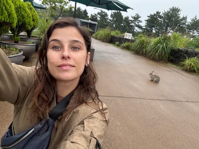 This is a selfie of a young woman taken outdoors in Seogwipo-si, South Korea. The image is oriented vertically, with the woman's face close to the bottom, looking upwards towards the camera. She has long, brown, slightly damp hair and striking blue eyes. She is wearing a light-colored, possibly khaki, jacket which appears speckled with water droplets. A dark blue bag with a silver zipper is slung across her body.
The background shows a paved walkway or path, with a small, light brown animal, possibly a rabbit or a rodent, standing on the path to the left of the woman. Beyond the path, there is lush green grass and foliage, with trees forming a dense canopy. In the distance, some structures and green umbrellas are visible. The overall atmosphere suggests an overcast or rainy day, as indicated by the dampness on the woman's hair and jacket, and the muted lighting. There is a white sign with text in Korean visible among the greenery, partially obscured by the foliage. The presence of potted plants and umbrellas suggests an outdoor recreational area, possibly a park or a garden. The woman's expression appears neutral.
