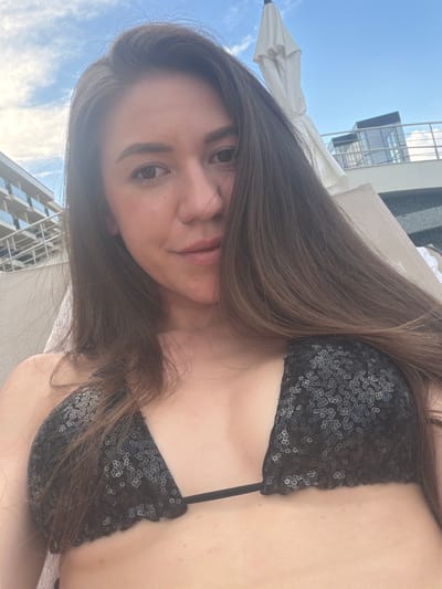 A young woman is lying down outdoors, wearing a black sequined bikini top. Her long brown hair is spread out around her. The background shows a bright blue sky with some clouds, the top of a modern building with geometric windows, and a white umbrella. The scene appears to be a sunny day, likely during daylight hours. The woman is looking towards the camera, and her expression is neutral or slightly contemplative. The overall setting suggests a resort or a rooftop area, possibly in Hisarya, Bulgaria. There is no visible text in the image.