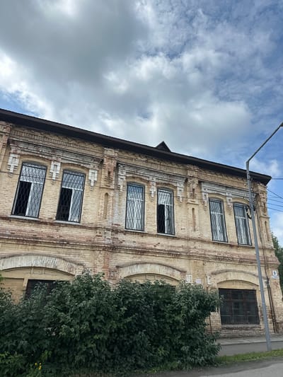 The image shows the facade of a two-story brick building in Nogornyi, Russia. The building has a weathered appearance with peeling paint and bricks showing signs of age. Several windows on the upper floor are visible, some with black metal security bars. The lower part of the building is partially obscured by dense green bushes. A street lamp and a sign with the number "9" are visible on the right side of the building. The sky above is cloudy.