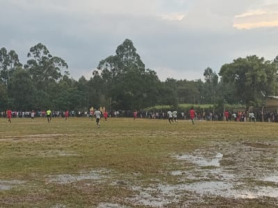 A soccer match is underway on a flooded field. Players wearing red and white jerseys are scattered across the field. A large crowd of spectators lines the perimeter, watching the game. Tall trees surround the field, and a cloudy sky looms overhead. The foreground is dominated by muddy patches and puddles of water, reflecting the overcast sky.