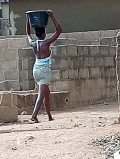 A woman carries a large, dark-colored basin on her head while walking in an outdoor, residential area in Gboko, Nigeria. She wears a short, patterned dress and sandals. The background consists of a low brick wall and dirt ground. The light indicates it is daytime.