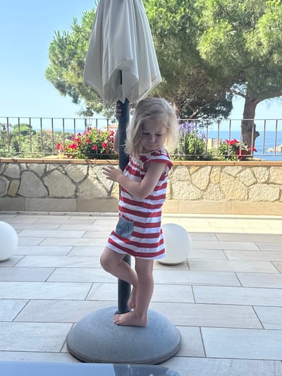 A young girl with blonde hair stands on the base of a patio umbrella, facing right. She is wearing a red and white striped dress with a grey pocket. Her left foot is placed on top of the umbrella base. The background shows a stone wall, a metal fence, flowering bushes, trees, and the sea under a clear blue sky. Two white spherical objects are visible on the tiled patio floor. The location is likely a terrace overlooking the Mediterranean Sea in Sant Feliu de Guíxols, Spain.