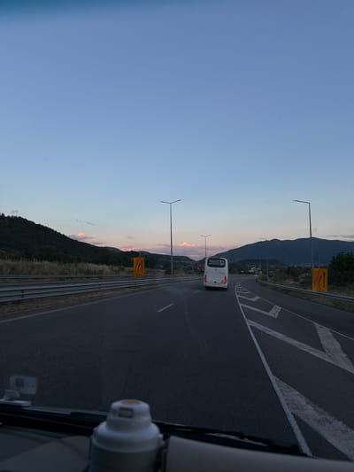 A clear, bright blue sky transitions into a soft pink and orange hue near the horizon, indicative of dusk or dawn. Below, a highway stretches into the distance, flanked by guardrails and sparse vegetation. Rolling hills covered in dark trees and dry grass line the left side of the road. On the right, mountains recede into the background. A white bus is visible in the distance on the highway, traveling away from the camera. Road signs indicating lane closures or merging traffic are present on both sides of the road. The foreground shows the dashboard of a vehicle, with a white object, possibly a GPS device or air freshener, prominently displayed. The overall scene is one of a journey on an open road at twilight, with natural landscapes surrounding the infrastructure. The image location is on the highway A3 near Simitli, Bulgaria.