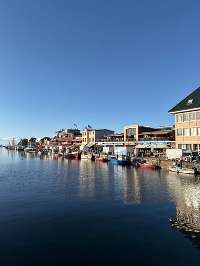 This is a horizontal, eye-level shot of the Warnow River in Rostock, Germany, specifically the harbor area. The sky is a clear, bright blue, with a few faint clouds on the far left. Along the riverbank, there is a row of buildings with shops and restaurants, many with outdoor seating covered by red awnings. Several boats are docked along the pier, including fishing boats and smaller pleasure craft. The buildings are reflected in the calm water of the river, creating a shimmering effect. The scene appears to be set in the late afternoon, as evidenced by the warm light and long shadows. There are a few people visible walking along the pier or sitting at the outdoor cafes, but the overall atmosphere is peaceful and serene. The architecture of the buildings suggests a maritime theme, with some featuring nautical flags and signage.
