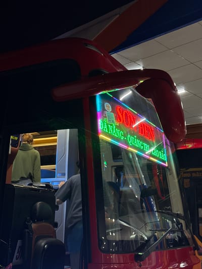 The image shows the interior of a bus, likely at a station or bus stop, in Diên Sanh Commune, Vietnam. The focus is on the front of the bus, with a vibrant LED sign displaying text in Vietnamese. The sign is illuminated in multiple colors, including red, green, and pink, and reads "SON TUAN" at the top, with "ĐÀ NẴNG - QUẢNG TỊ." below it. The words "TÍCH LỮ" appear to be partially visible on the right side of the sign.

Inside the bus, a person wearing a green hooded sweatshirt is visible from the back, standing near a grey machine that resembles a ticket dispenser or a payment terminal. Another person, wearing a dark t-shirt, is partially visible on the right, seemingly interacting with the same machine. The bus interior features dark seats, some of which are visible in the foreground. The overall environment appears to be dimly lit, with the bright LED sign being the primary light source in the immediate area. Reflections on the bus windshield suggest an indoor or covered area, possibly a terminal or garage. In the background, there are shelves and a dark ceiling. The side of the bus shows a yellow sign with red text and a number "5" inside a circle.